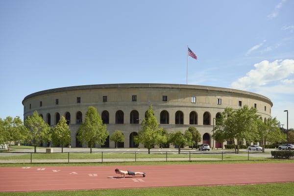 A wide view of the exterior of a large, circular stadium (likely Harvard Stadium) with Roman-style arches, a red running track in the foreground where a person is doing a push-up, and an American flag waving on top.