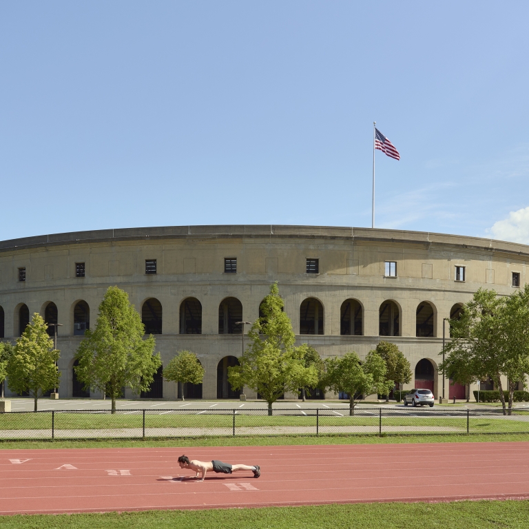 A wide view of the exterior of a large, circular stadium (likely Harvard Stadium) with Roman-style arches, a red running track in the foreground where a person is doing a push-up, and an American flag waving on top.