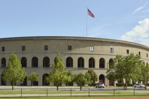 A wide view of the exterior of a large, circular stadium (likely Harvard Stadium) with Roman-style arches, a red running track in the foreground where a person is doing a push-up, and an American flag waving on top.
