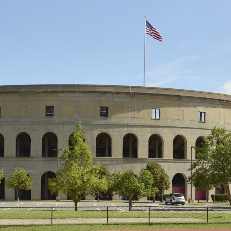 A wide view of the exterior of a large, circular stadium (likely Harvard Stadium) with Roman-style arches, a red running track in the foreground where a person is doing a push-up, and an American flag waving on top.