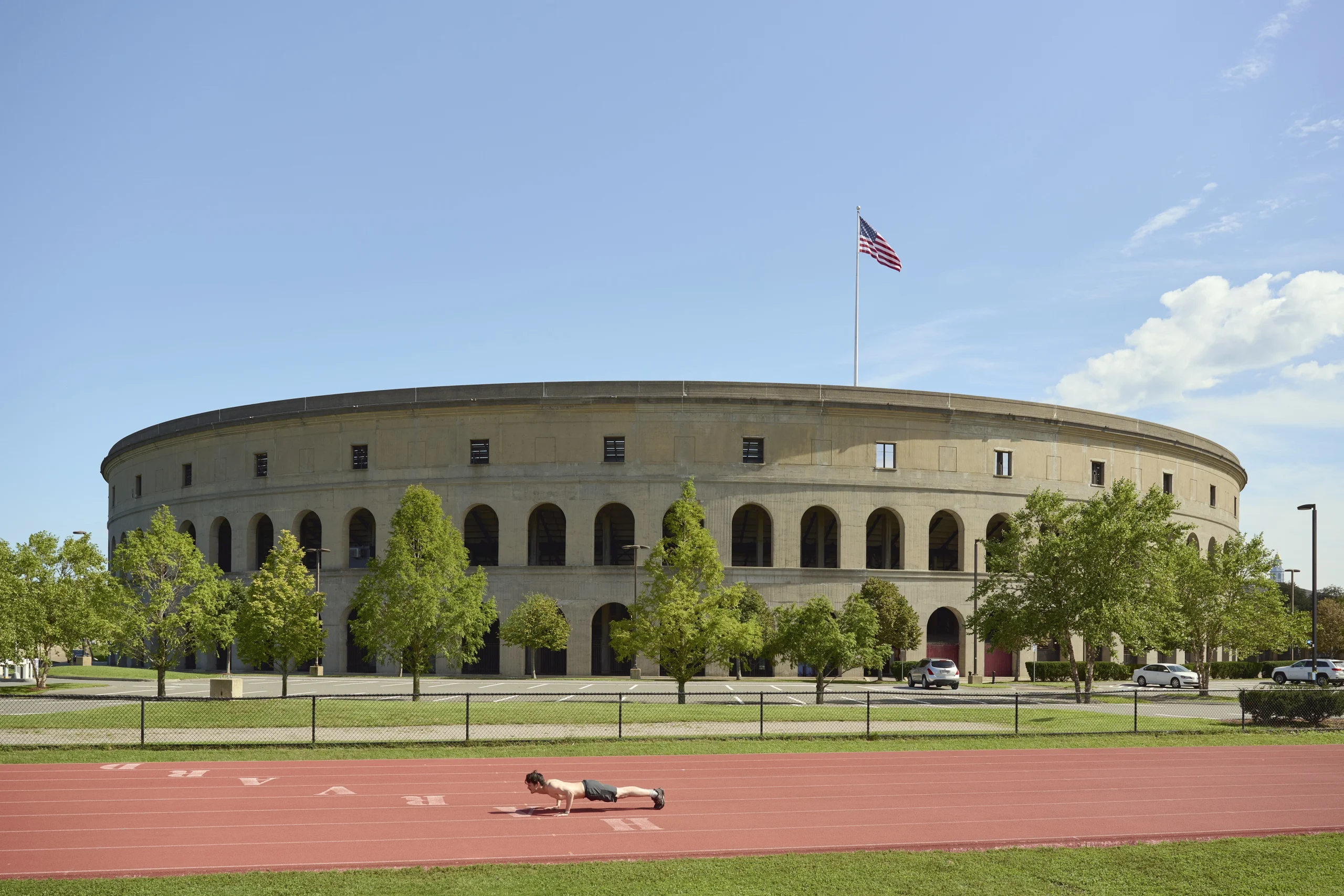 A wide view of the exterior of a large, circular stadium (likely Harvard Stadium) with Roman-style arches, a red running track in the foreground where a person is doing a push-up, and an American flag waving on top.
