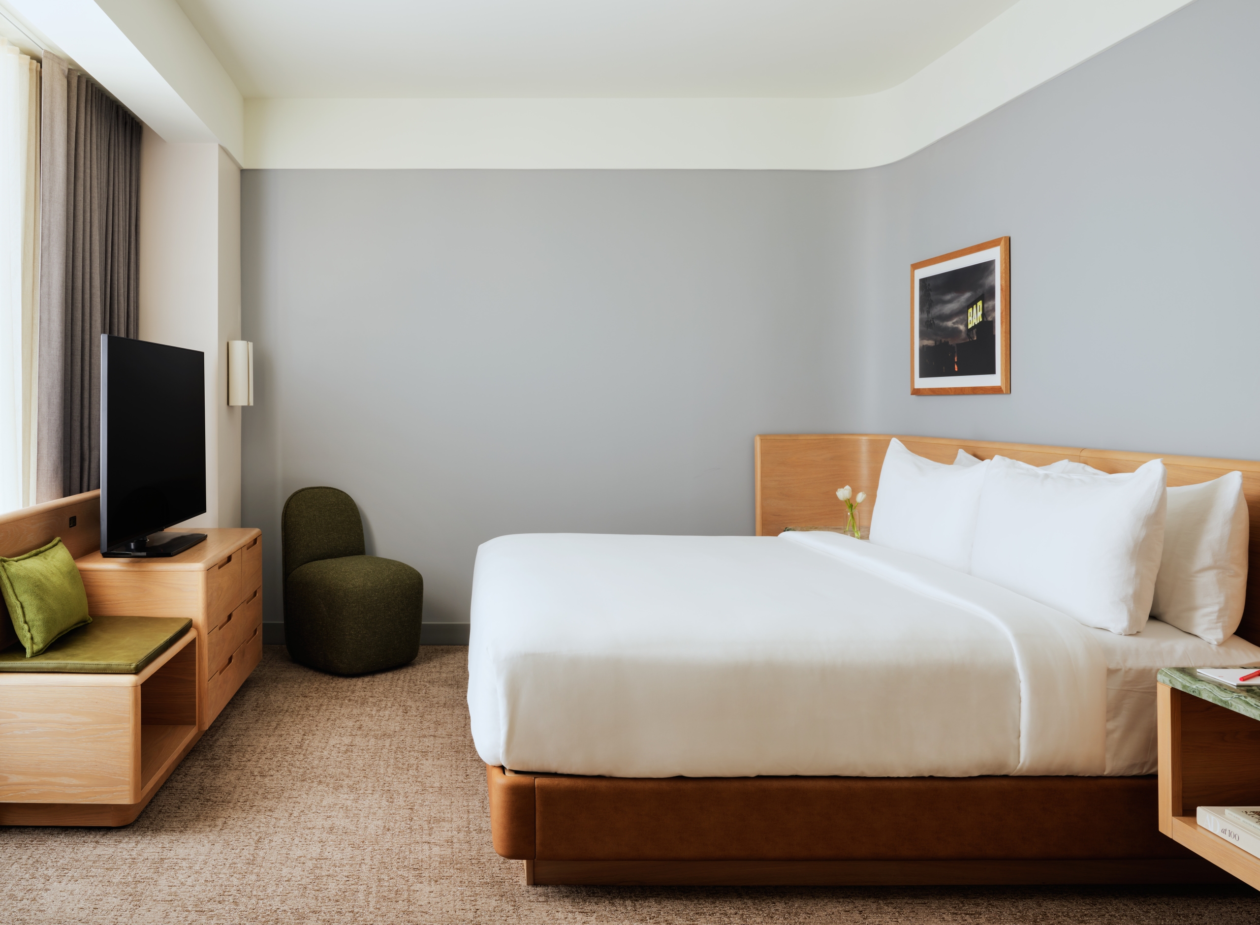 Hotel room with a large bed, white linens, wooden headboard and furniture, gray walls, a framed photo, and a TV on a wooden stand near a window with curtains.