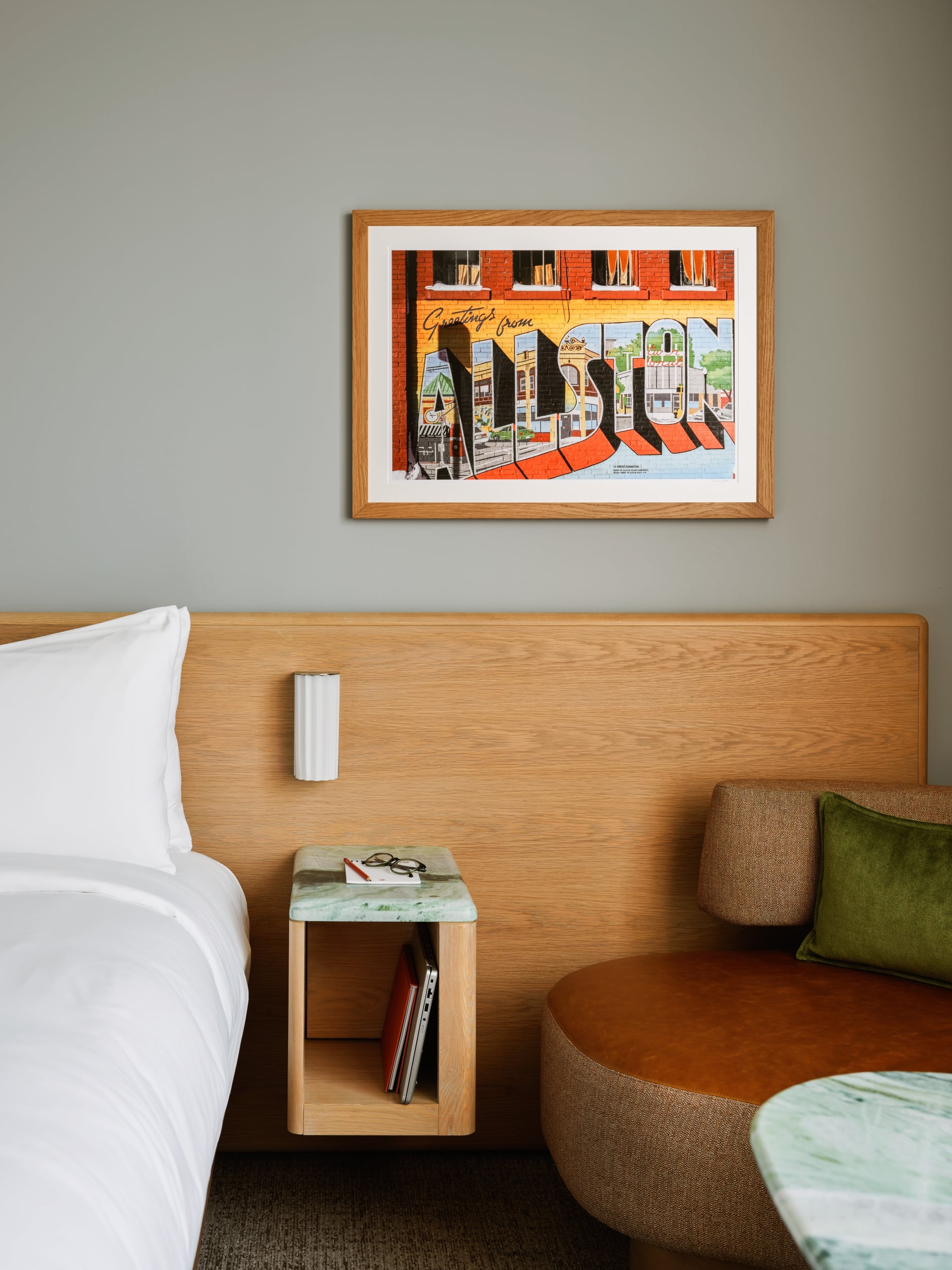 Framed colorful art print hanging above a wooden headboard in a hotel room. A nightstand with glasses and a pencil rests beside the bed.