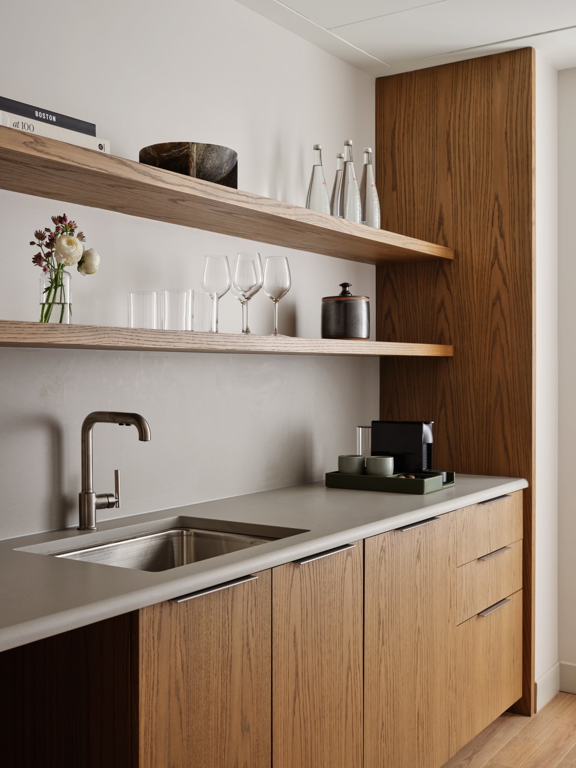 Minimalist kitchen with floating wooden shelves holding glassware, bottles, and a small vase of flowers above a light countertop and sink.