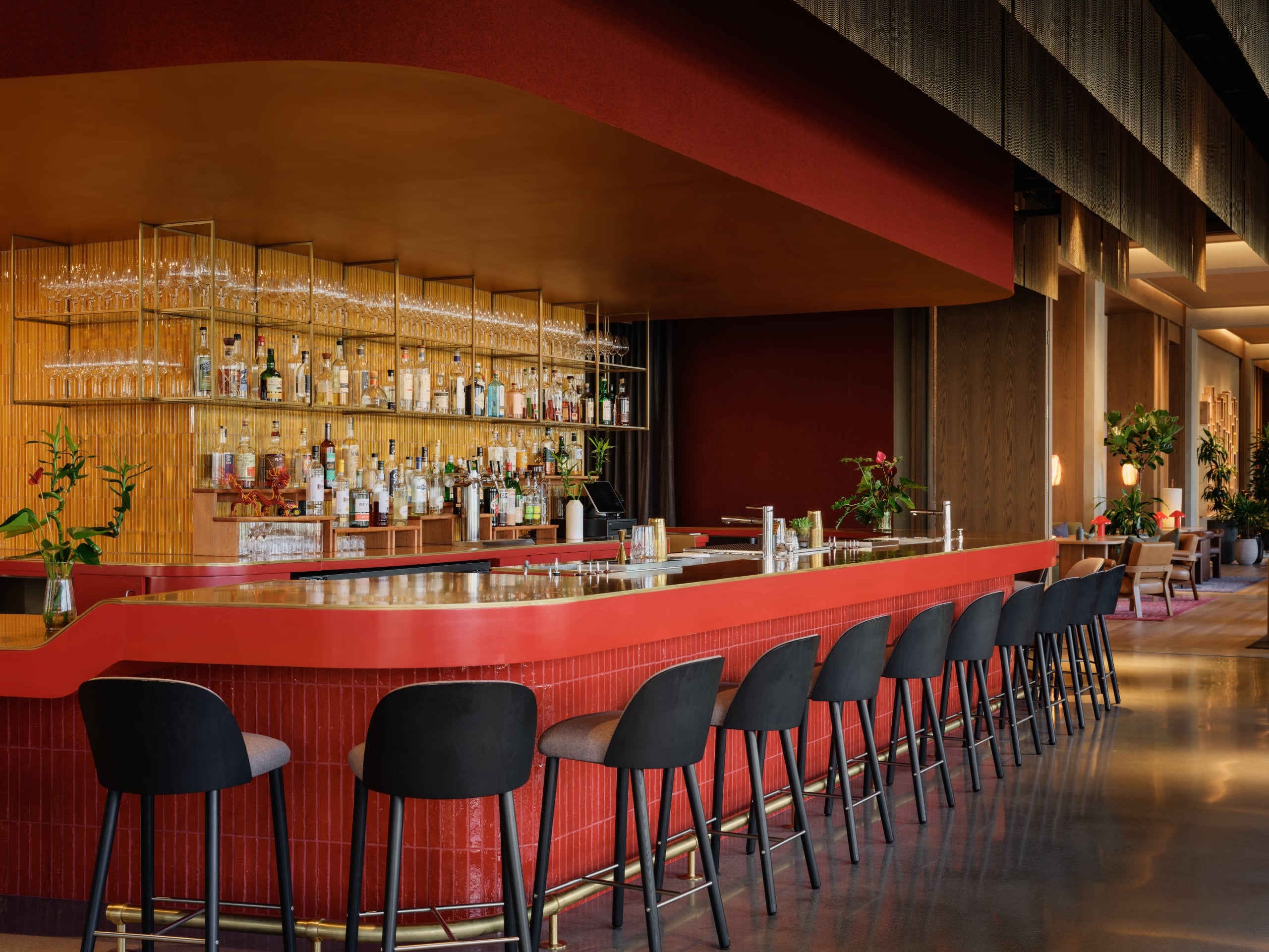 A bold red tiled bar with black stools faces a backlit shelf of spirits and hanging glassware, set within a warmly lit interior.