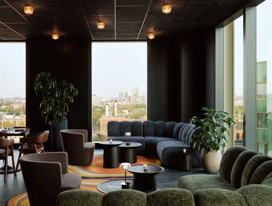 A darkly lit rooftop lounge with curved velvet seating, black tables, and floor-to-ceiling windows overlooking a city skyline.