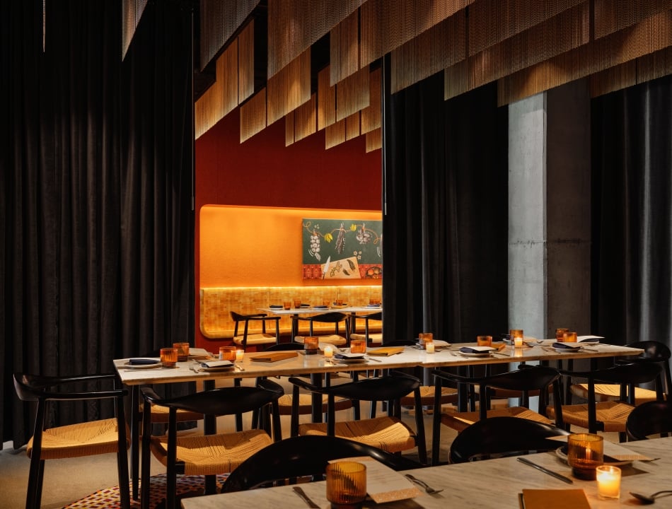 A restaurant dining room with marble tables lit by candlelight, woven chairs, dark draping, and a glowing amber banquette beneath artwork.