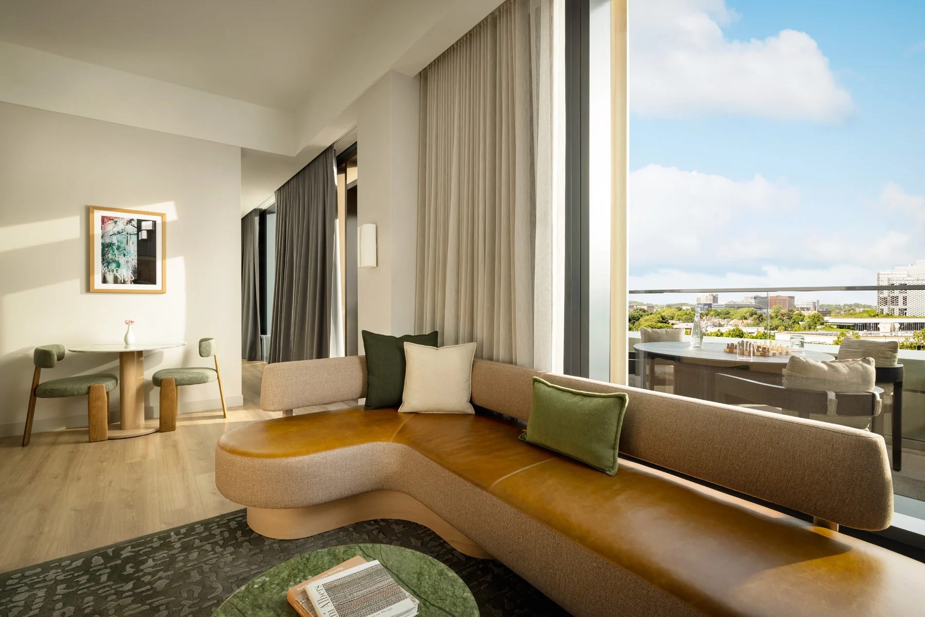 Bright living room with a curved tan sofa, green cushions, and floor-to-ceiling windows looking out to the city skyline.