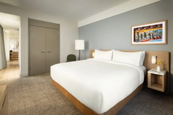 Hotel bedroom with a large white bed, gray wall, framed colorful mural, and nightstand with a lamp on the right.