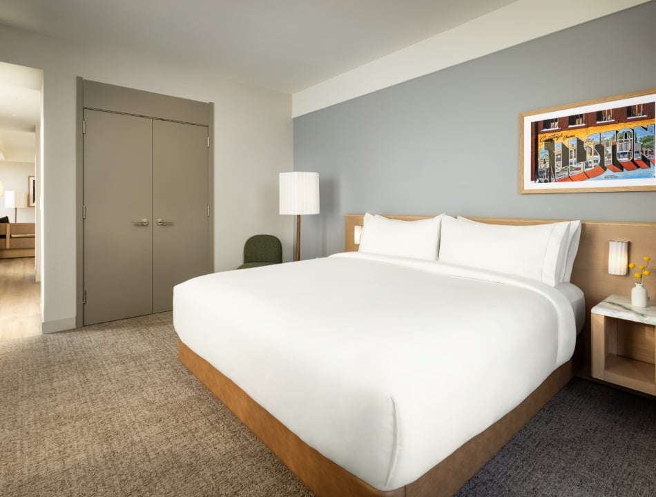 Hotel bedroom with a large white bed, gray wall, framed colorful mural, and nightstand with a lamp on the right.