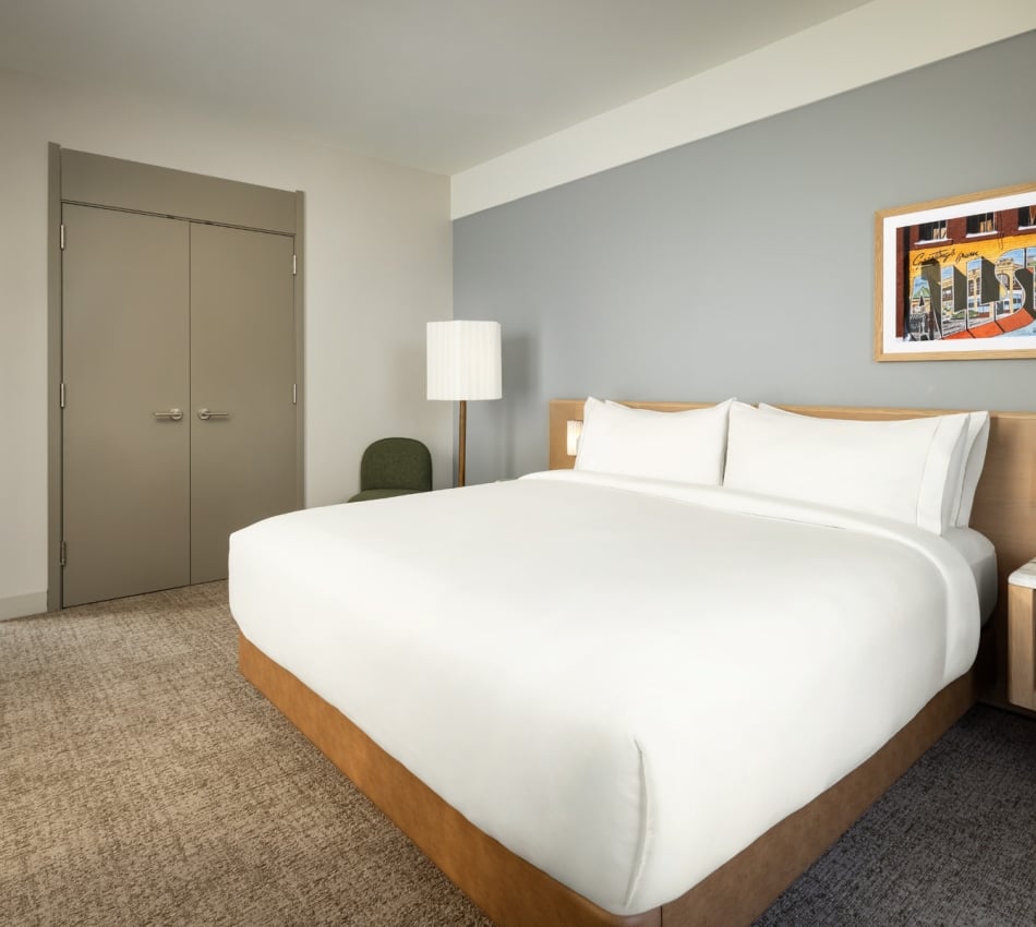 Hotel bedroom with a large white bed, gray wall, framed colorful mural, and nightstand with a lamp on the right.