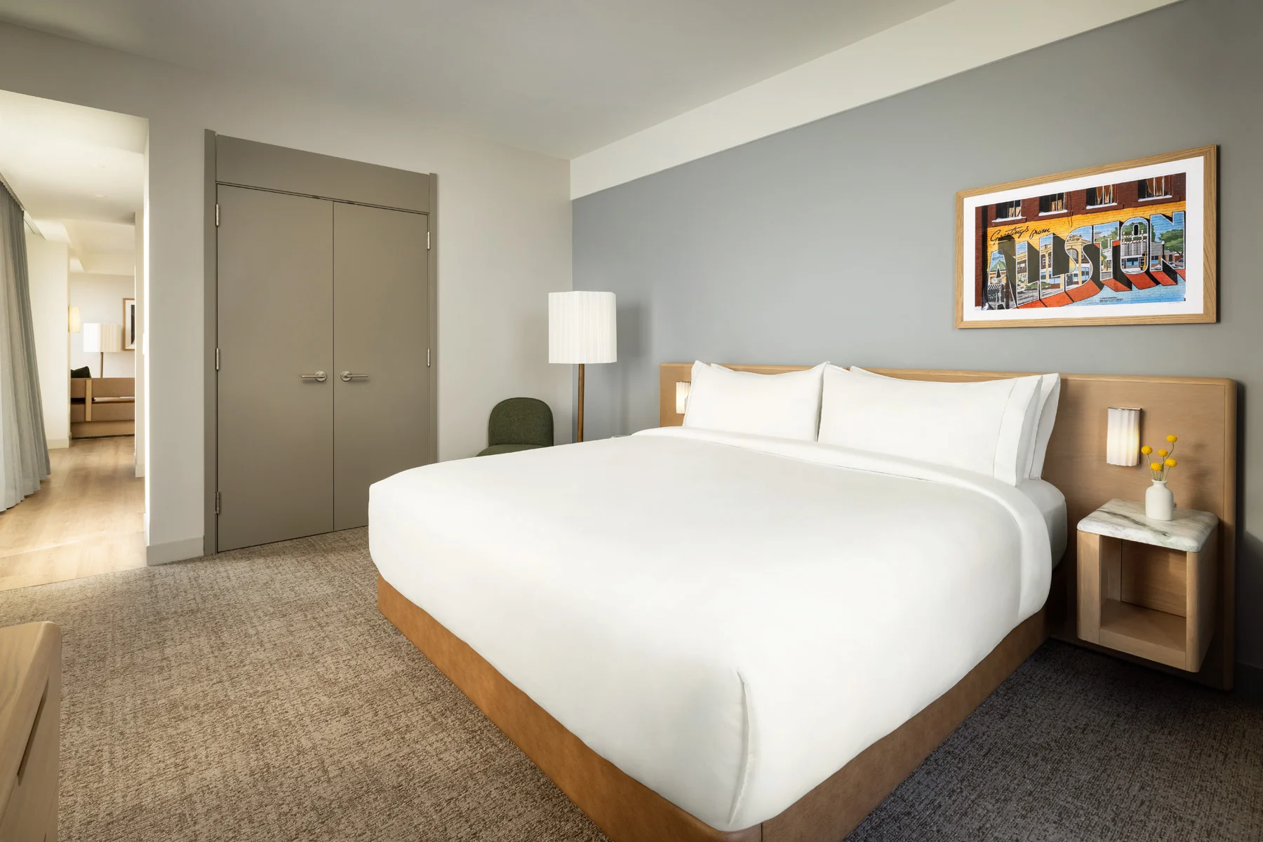 Hotel bedroom with a large white bed, gray wall, framed colorful mural, and nightstand with a lamp on the right.