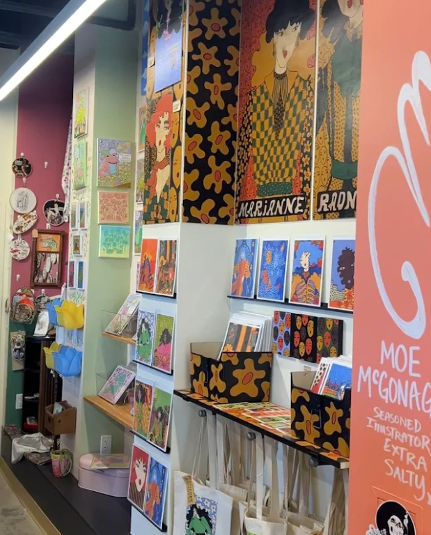 A colorful corner inside the Brighton Emporium, showcasing local artwork, prints, cards, and canvas tote bags on walls painted in green and magenta.