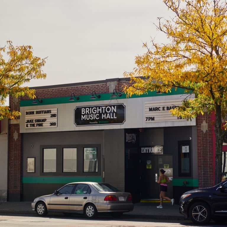 The exterior of the Brighton Music Hall, a low-rise brick venue with a marquee displaying the venue name and show listings, seen on a sunny day with yellow-leafed trees and cars parked on the street.