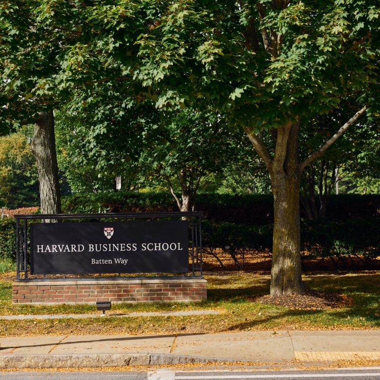A close-up of the Harvard Business School sign on a low brick wall, partially framed by the lush green leaves of a large tree. The sign reads 