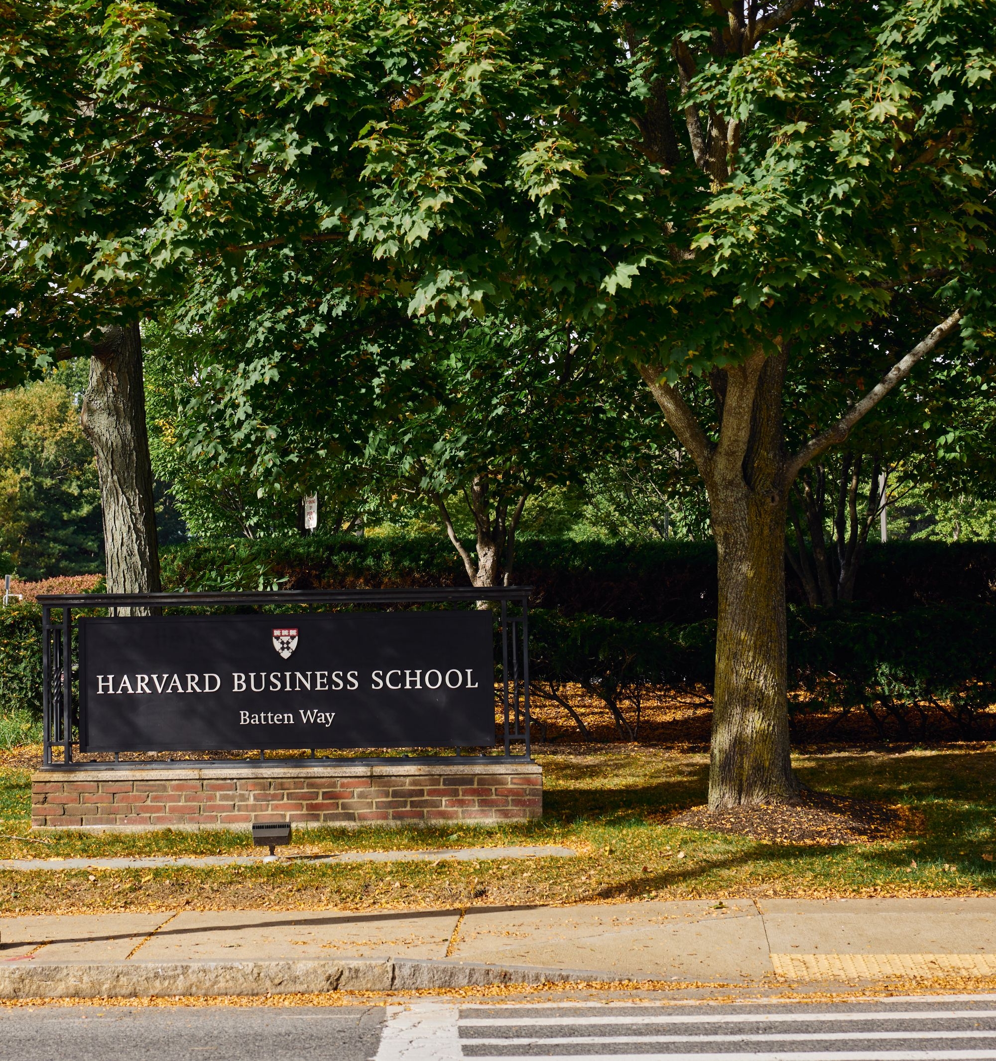 A close-up of the Harvard Business School sign on a low brick wall, partially framed by the lush green leaves of a large tree. The sign reads
