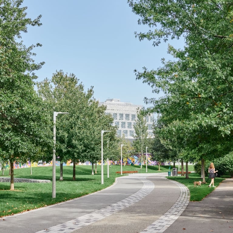 A scenic, paved walking path curving through Honan-Park, lined with young trees and grass. A person is walking a small dog on the right, with a large, modern white building visible in the distance.