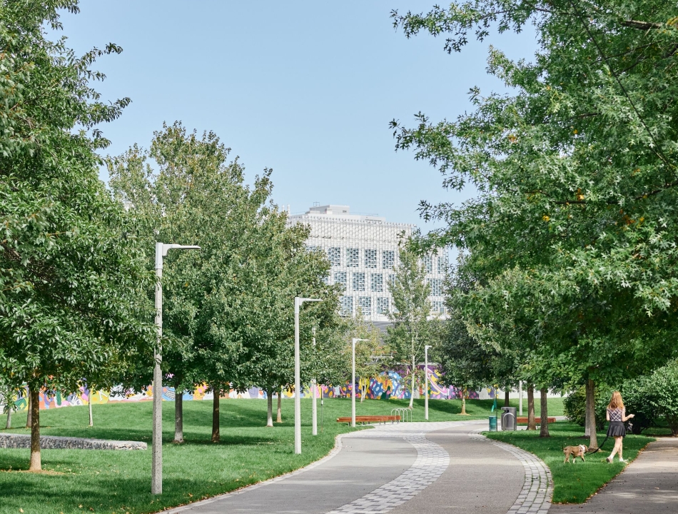 A scenic, paved walking path curving through Honan-Park, lined with young trees and grass. A person is walking a small dog on the right, with a large, modern white building visible in the distance.