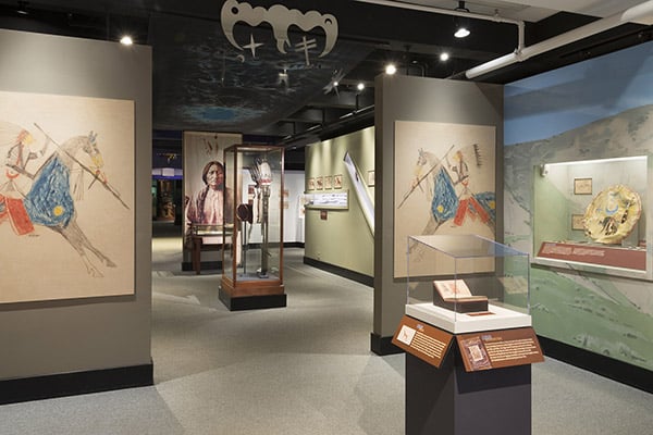 An interior exhibit hall at the Peabody Museum of Archaeology and Ethnology, featuring large painted canvas illustrations of Native American figures on horseback, displayed between glass cases of artifacts.