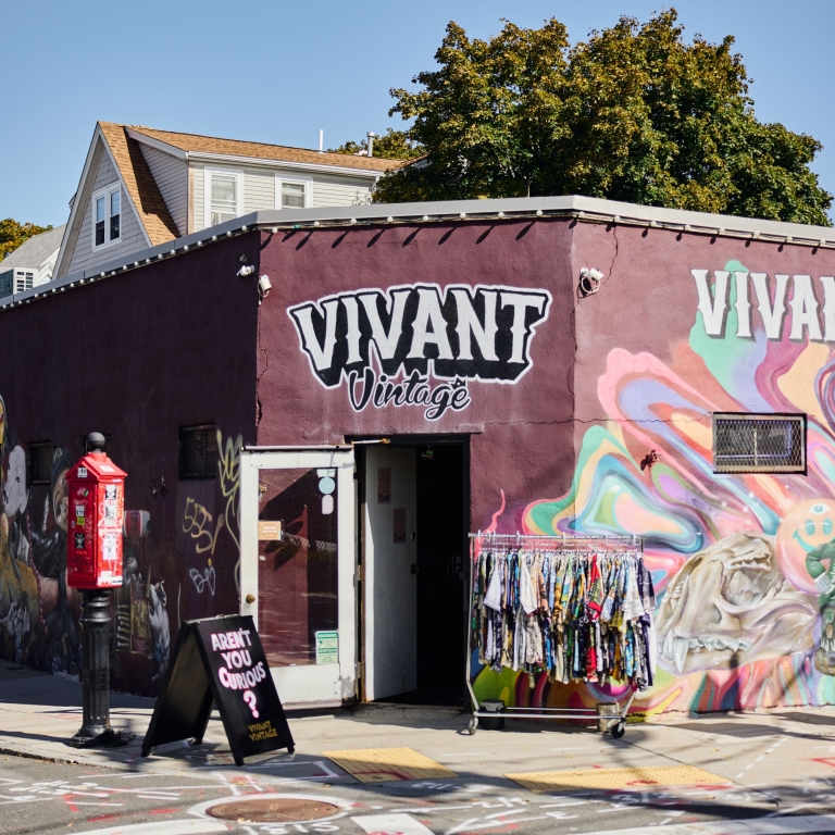 The exterior of a Vivant Vintage store building with a dark purple facade and a bold white logo. The walls are covered in colorful street art and murals, with a rack of clothing visible near the open doorway.