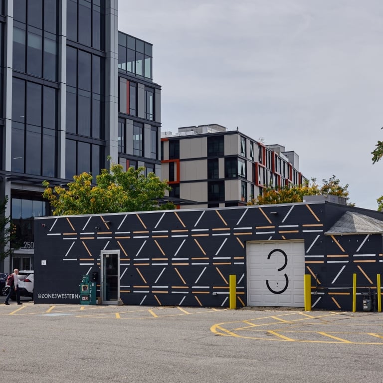 The exterior of Zone3, a single-story brick building painted black with a striking white and gold geometric pattern, sitting in a paved parking lot next to a modern, glass skyscraper. A white garage door features a stylized number '3'.