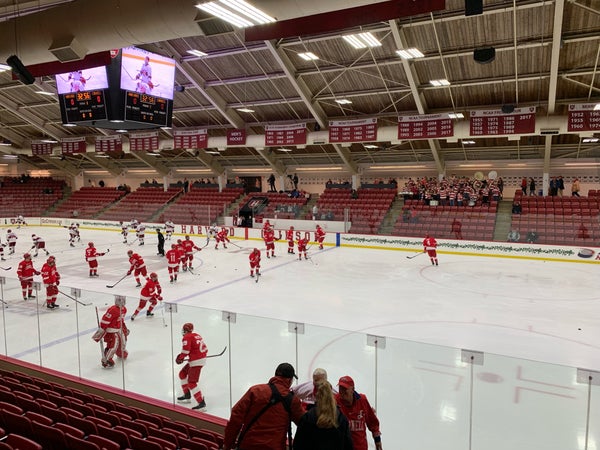 The interior of the Bright-Landry Hockey Center arena, showing a Harvard Crimson hockey team practicing on the ice, surrounded by empty red tiered seating.
