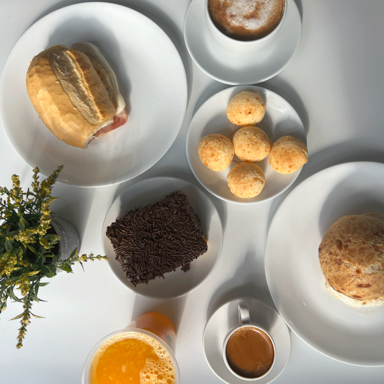 A bright, overhead view of a selection of Brazilian bakery items on white plates, including pão de queijo (cheese bread), a ham and cheese sandwich, a chocolate brigadeiro cake, and two cups of coffee.