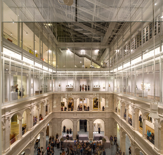 An interior view of the Harvard Art Museums' central courtyard, featuring three tiers of glass-enclosed galleries surrounding a traditional, arched lower level where a crowd is gathered.