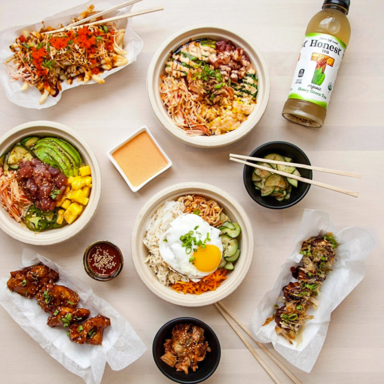 An overhead arrangement of several Korean-inspired food items from Kimchipapi, including poke bowls, Korean fried chicken wings, loaded fries, and a bottle of iced tea, all on a light wooden table.