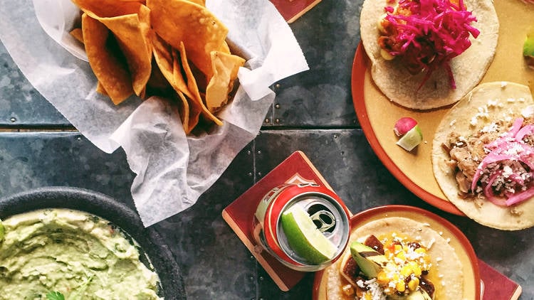 A rustic, overhead close-up of a spread of tacos and guacamole on a dark metal table, including a basket of tortilla chips and a beer with a lime slice.