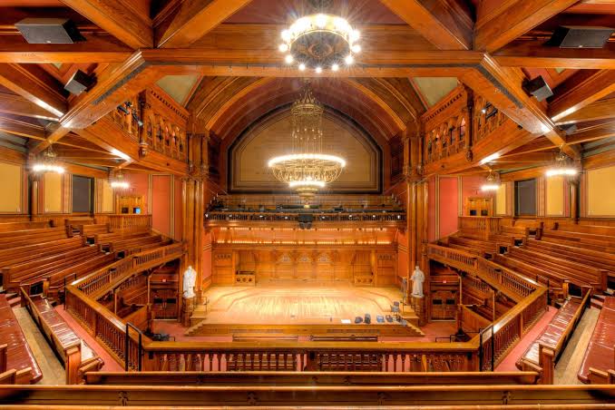 The grand interior of Sanders Theater at Harvard University, featuring tiered seating on multiple levels and an ornate, high ceiling made of dark carved wood, with a large chandelier hanging above the stage.