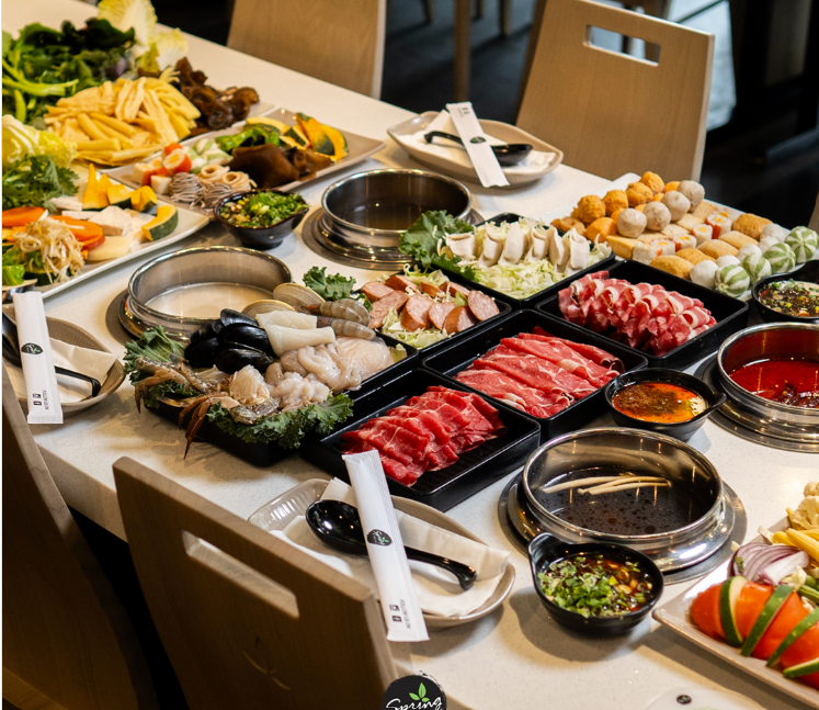 A wide table view filled with trays of ingredients for Shabu Shabu or hot pot, including thinly sliced raw meats, seafood, fresh vegetables, mushrooms, and various dipping sauces.