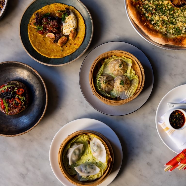An overhead flat lay of a variety of colorful dishes from Waypoint, including dim sum, pizza, and a small appetizer, arranged on a marble tabletop with a set of red chopsticks.