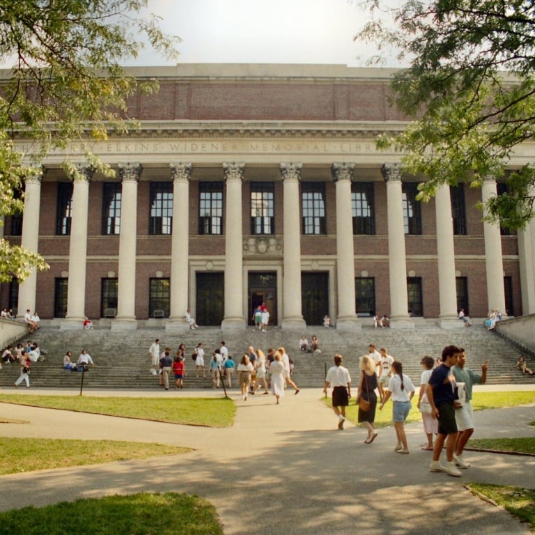 The massive, imposing Widener Memorial Library building at Harvard, featuring a brick facade and tall white columns. Students are walking up and sitting on the wide stone steps on a sunny day.
