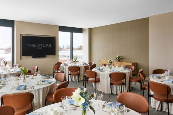 Banquet hall with round tables set for a formal dinner, floral centerpieces, glassware, and a wall-mounted TV reading 'THE ATLAS HOTEL'.