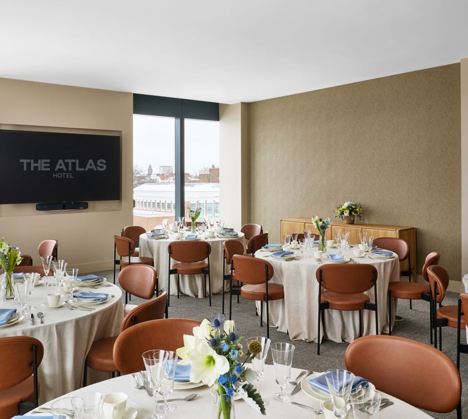 Banquet hall with round tables set for a formal dinner, floral centerpieces, glassware, and a wall-mounted TV reading 'THE ATLAS HOTEL'.