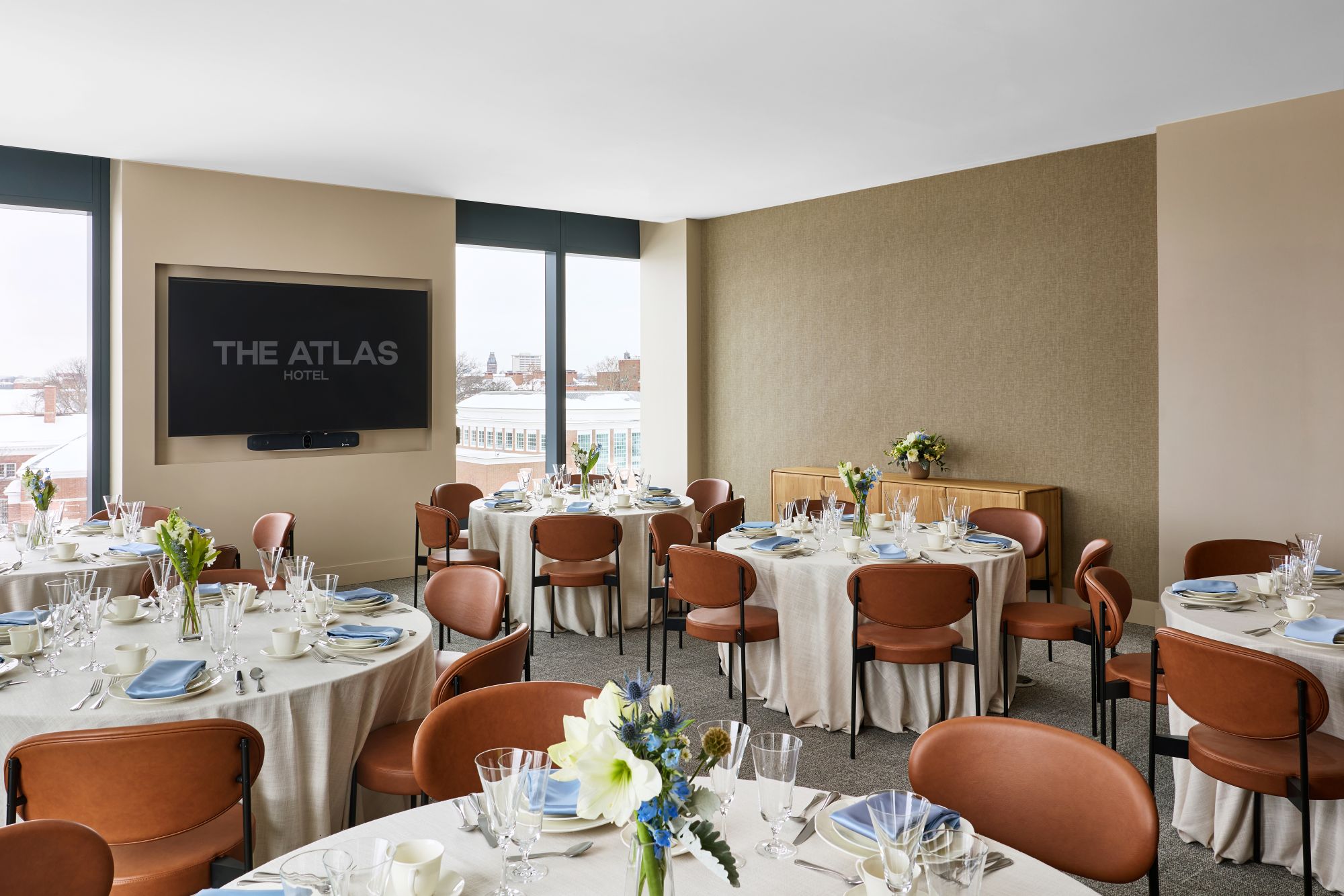 Banquet hall with round tables set for a formal dinner, floral centerpieces, glassware, and a wall-mounted TV reading 'THE ATLAS HOTEL'.