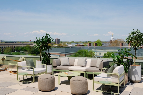 Rooftop lounge with beige sofas, green metal frames, and potted plants against a cityscape and blue sky.