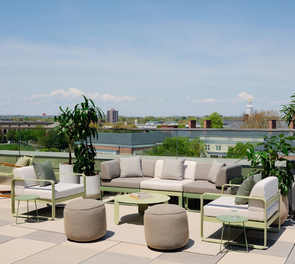 Rooftop lounge with beige sofas, green metal frames, and potted plants against a cityscape and blue sky.