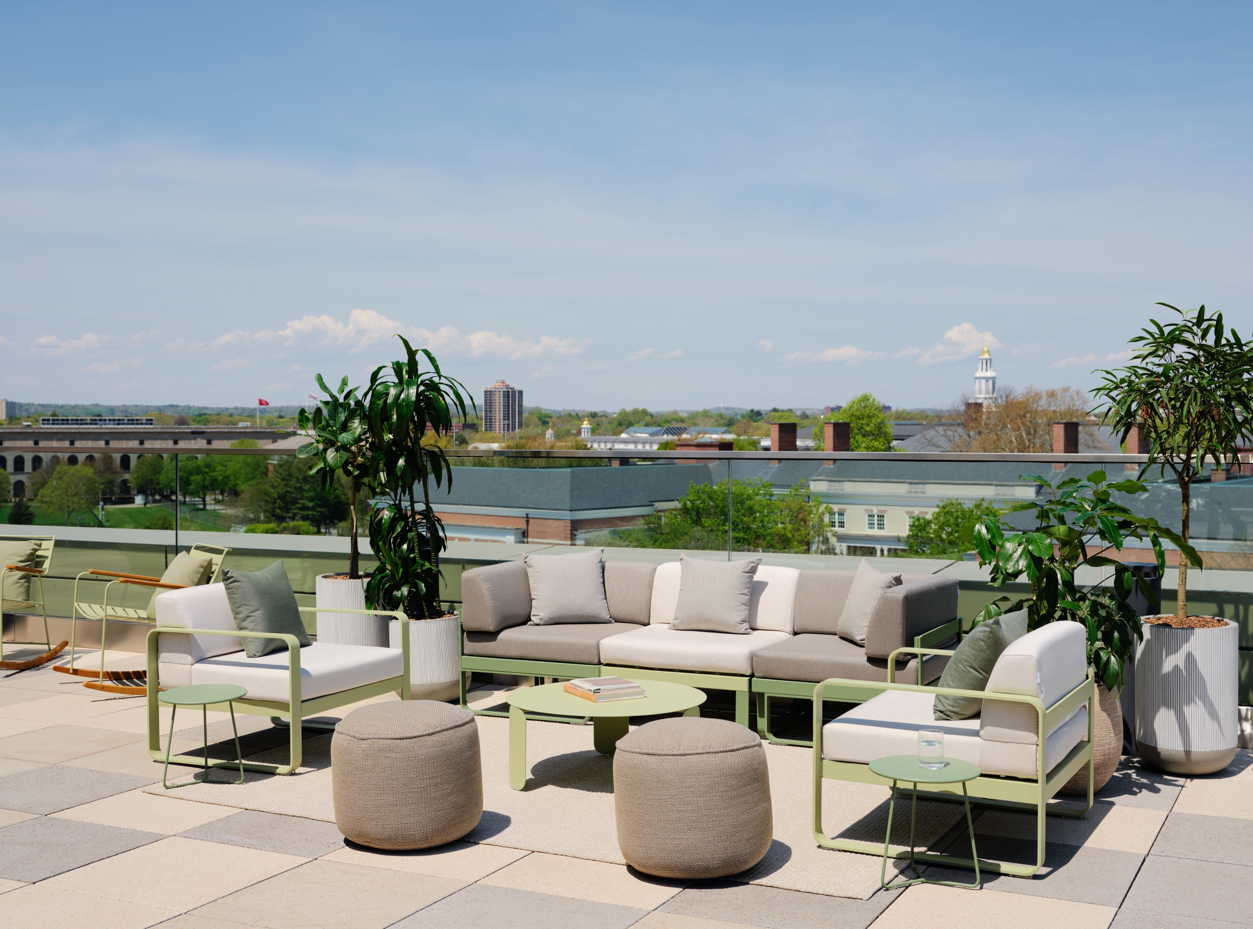 Rooftop lounge with beige sofas, green metal frames, and potted plants against a cityscape and blue sky.