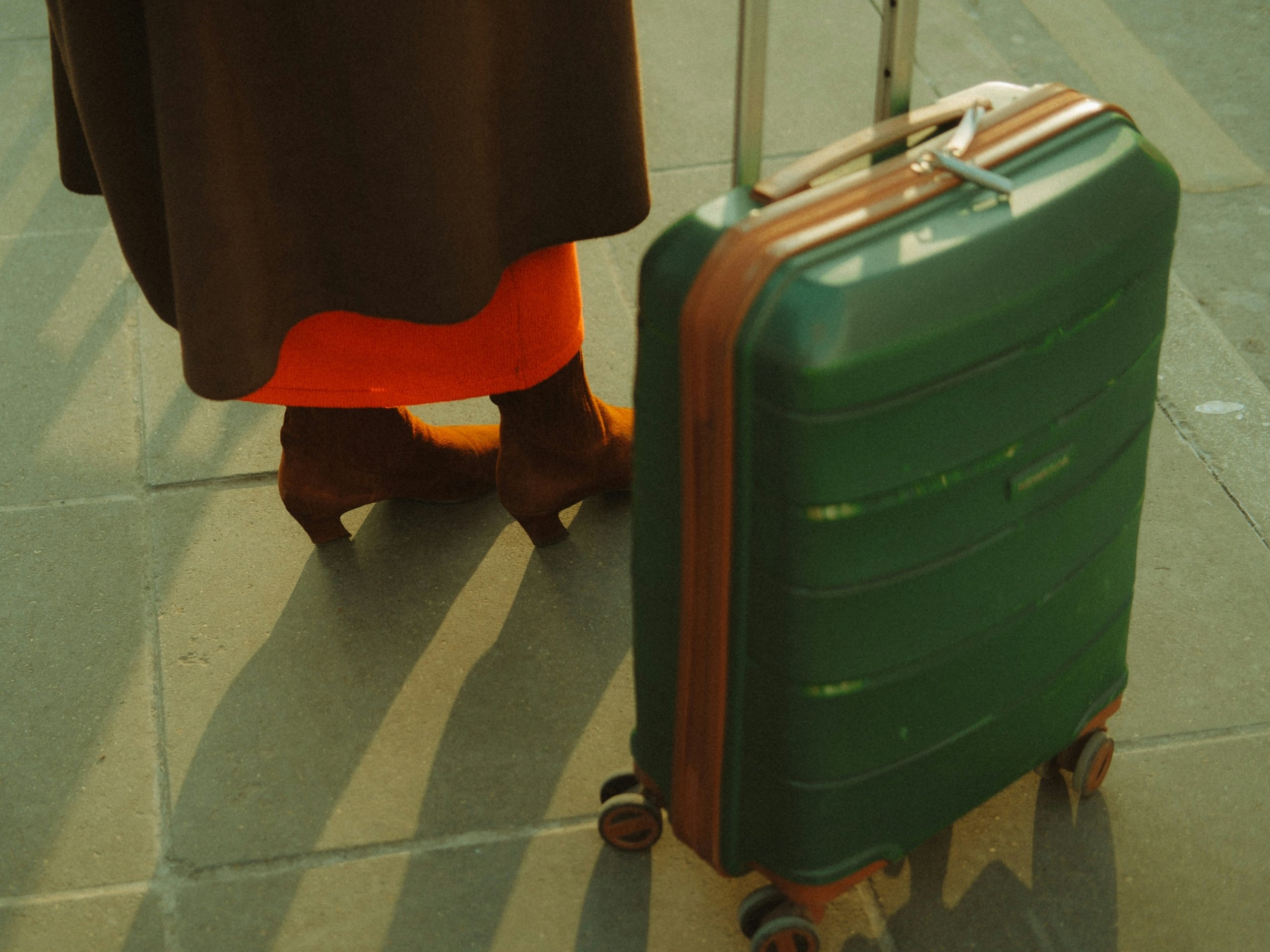 A close-up, low-angle shot of a person's legs, wearing a brown skirt, bright orange undergarment, and brown heeled boots, standing next to a green rolling suitcase with brown trim on a paved surface.