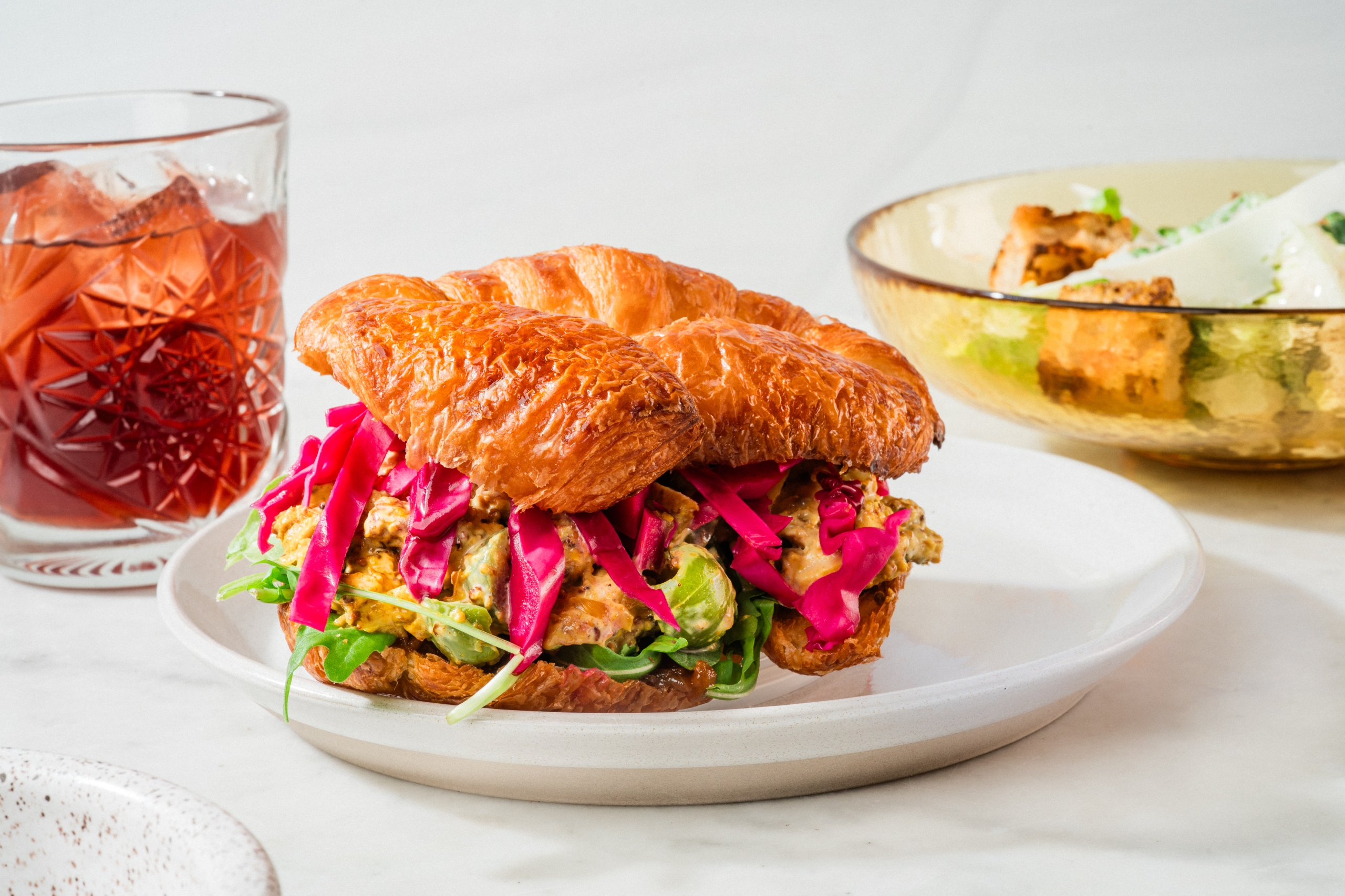A close-up of a large, golden-brown croissant sandwich overflowing with green chicken salad and bright pink pickled cabbage, served on a white plate next to a glass of iced red beverage.