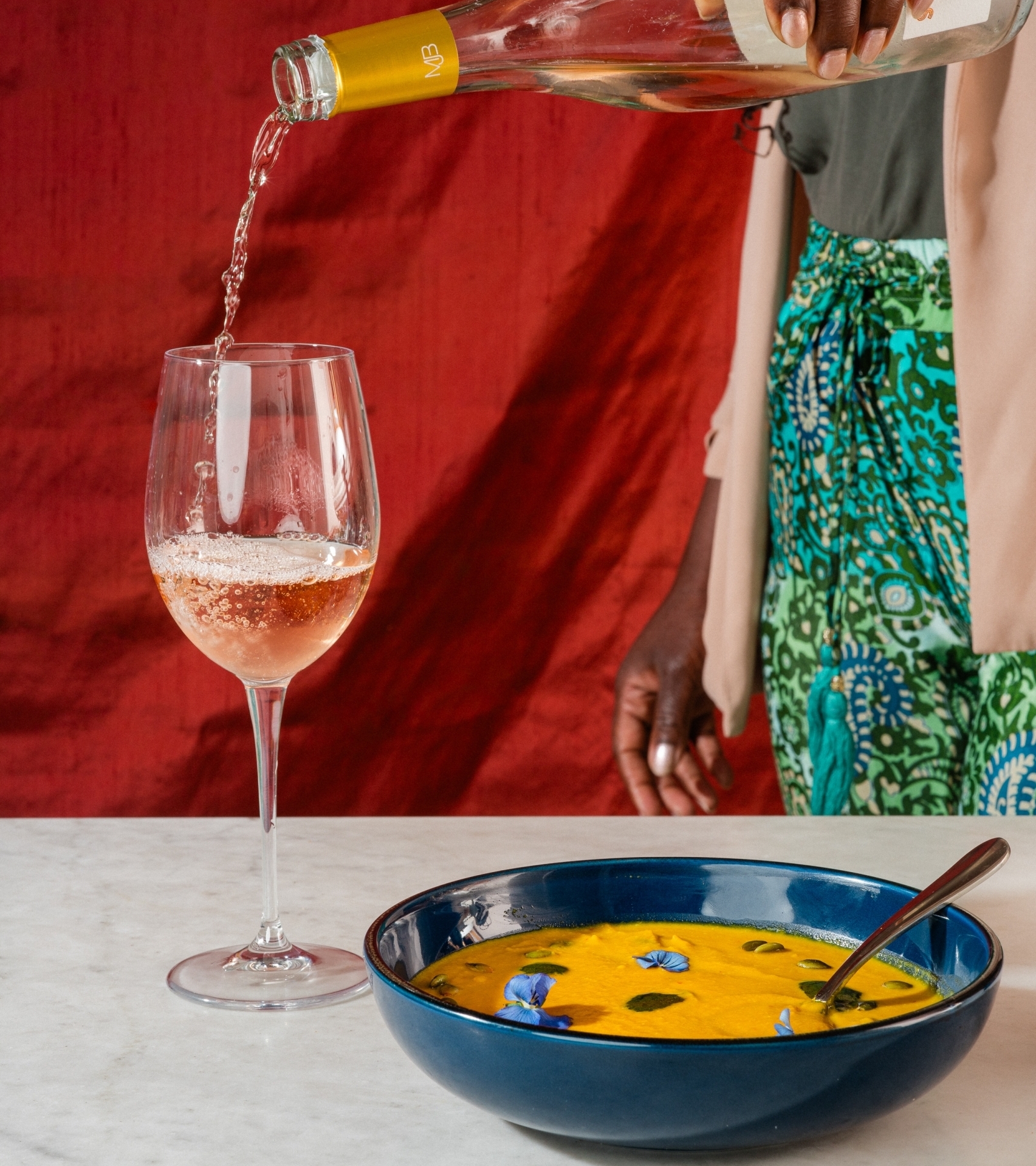 A person pours a glass of rosé wine next to a dark blue bowl filled with bright orange soup garnished with purple and white edible flowers, all set on a marble countertop.