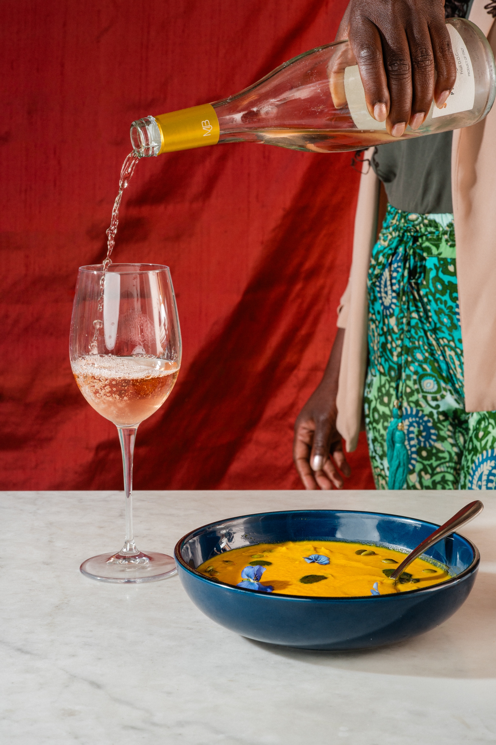 A person pours a glass of rosé wine next to a dark blue bowl filled with bright orange soup garnished with purple and white edible flowers, all set on a marble countertop.