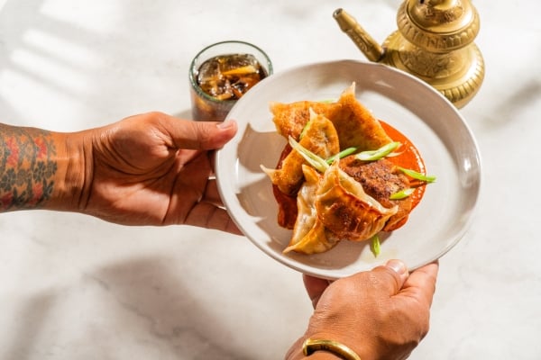 A tattooed person's hands hold a white plate with crispy dumplings resting on a bed of red sauce, garnished with scallions. A brass teapot and a glass of iced drink are visible in the background.