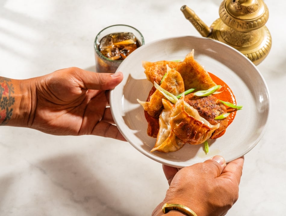 A tattooed person's hands hold a white plate with crispy dumplings resting on a bed of red sauce, garnished with scallions. A brass teapot and a glass of iced drink are visible in the background.