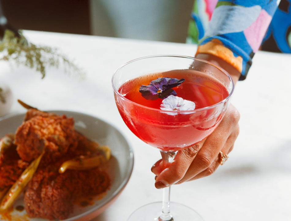 A person wearing a colorful floral dress holds a coupe glass filled with a bright pink cocktail garnished with edible flowers. A plate with fried food is blurred in the background.