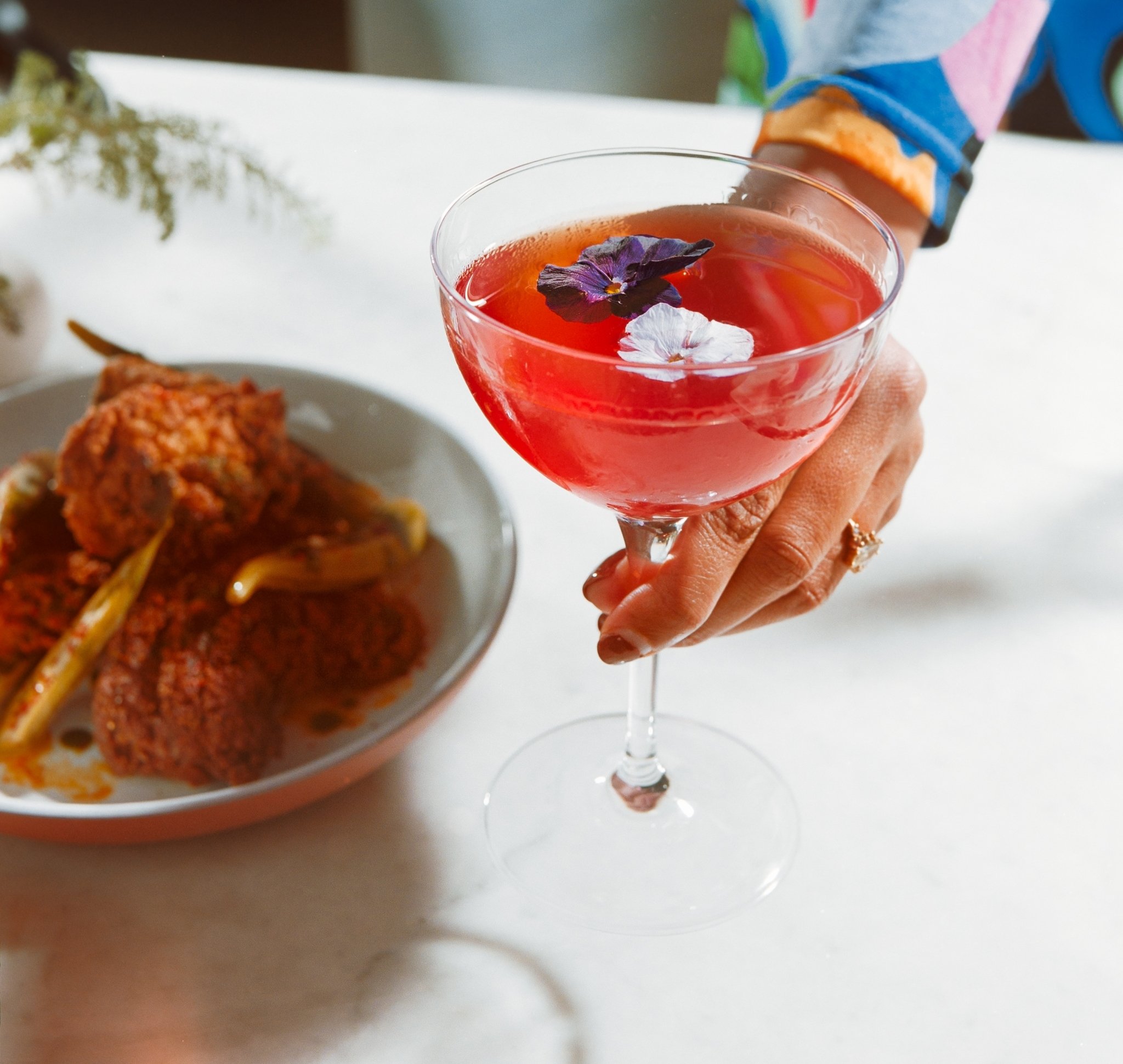 A person wearing a colorful floral dress holds a coupe glass filled with a bright pink cocktail garnished with edible flowers. A plate with fried food is blurred in the background.