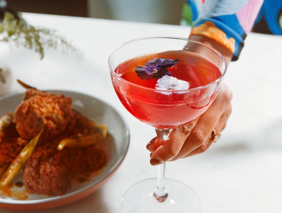 A person wearing a colorful floral dress holds a coupe glass filled with a bright pink cocktail garnished with edible flowers. A plate with fried food is blurred in the background.