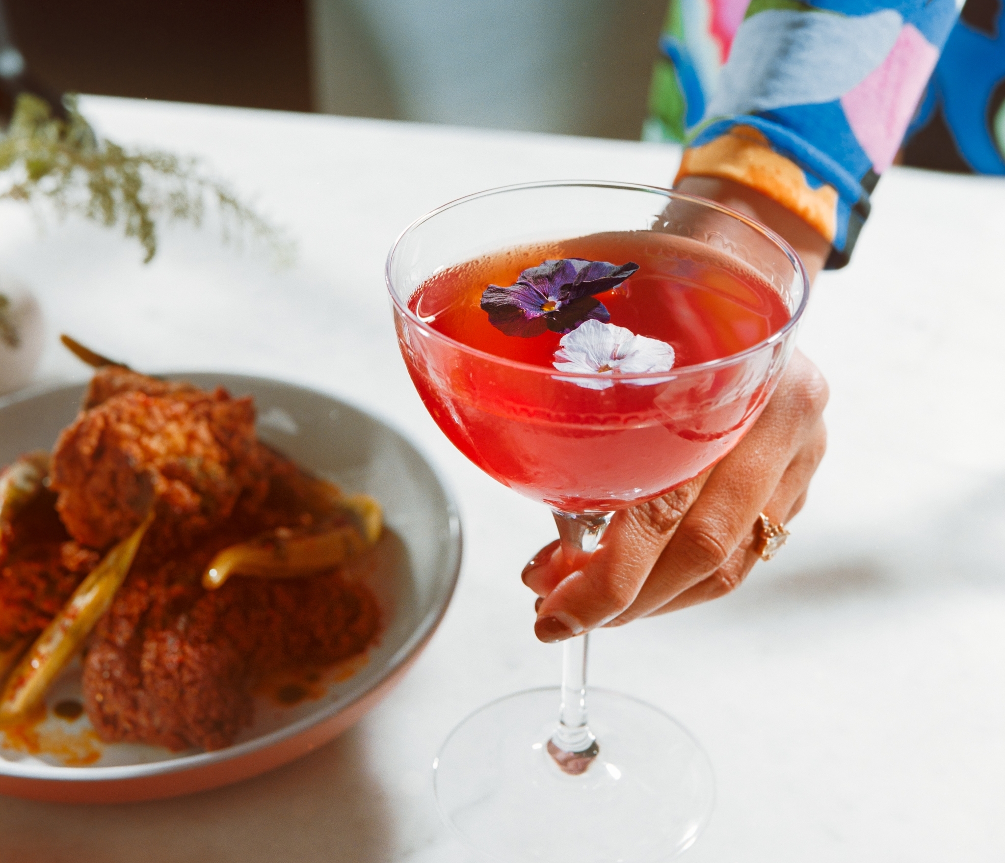 A person wearing a colorful floral dress holds a coupe glass filled with a bright pink cocktail garnished with edible flowers. A plate with fried food is blurred in the background.